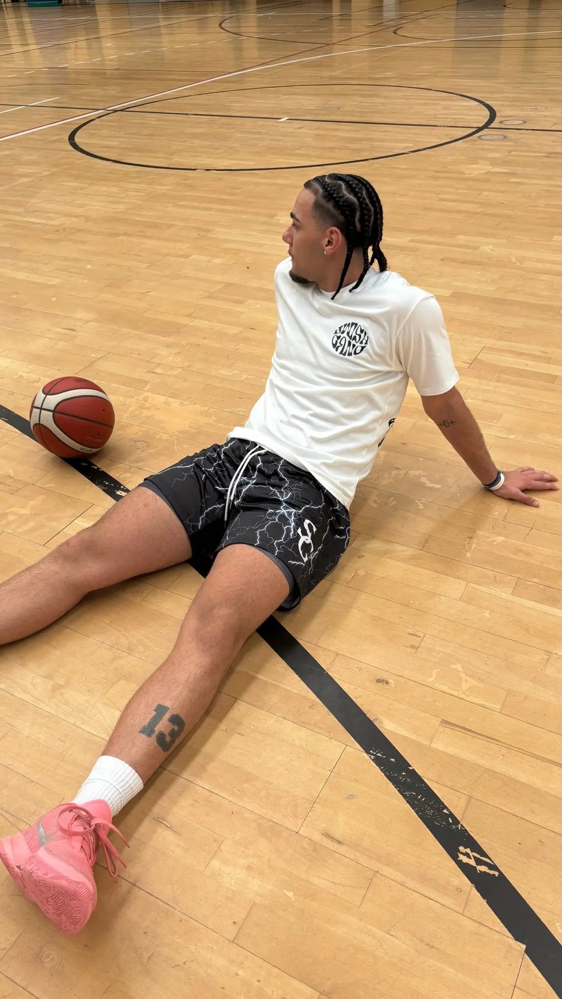A basketball player sitting on the floor of an indoor court, wearing black basketball shorts with a white lightning bolt pattern and a basketball beside him