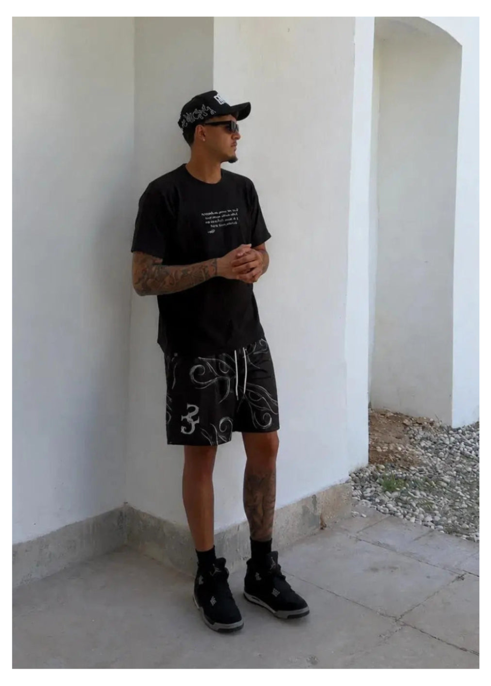 Lifestyle shot of a model wearing black printed basketball shorts, a black t-shirt, matching cap, and sneakers while posing outdoors against a white wall.