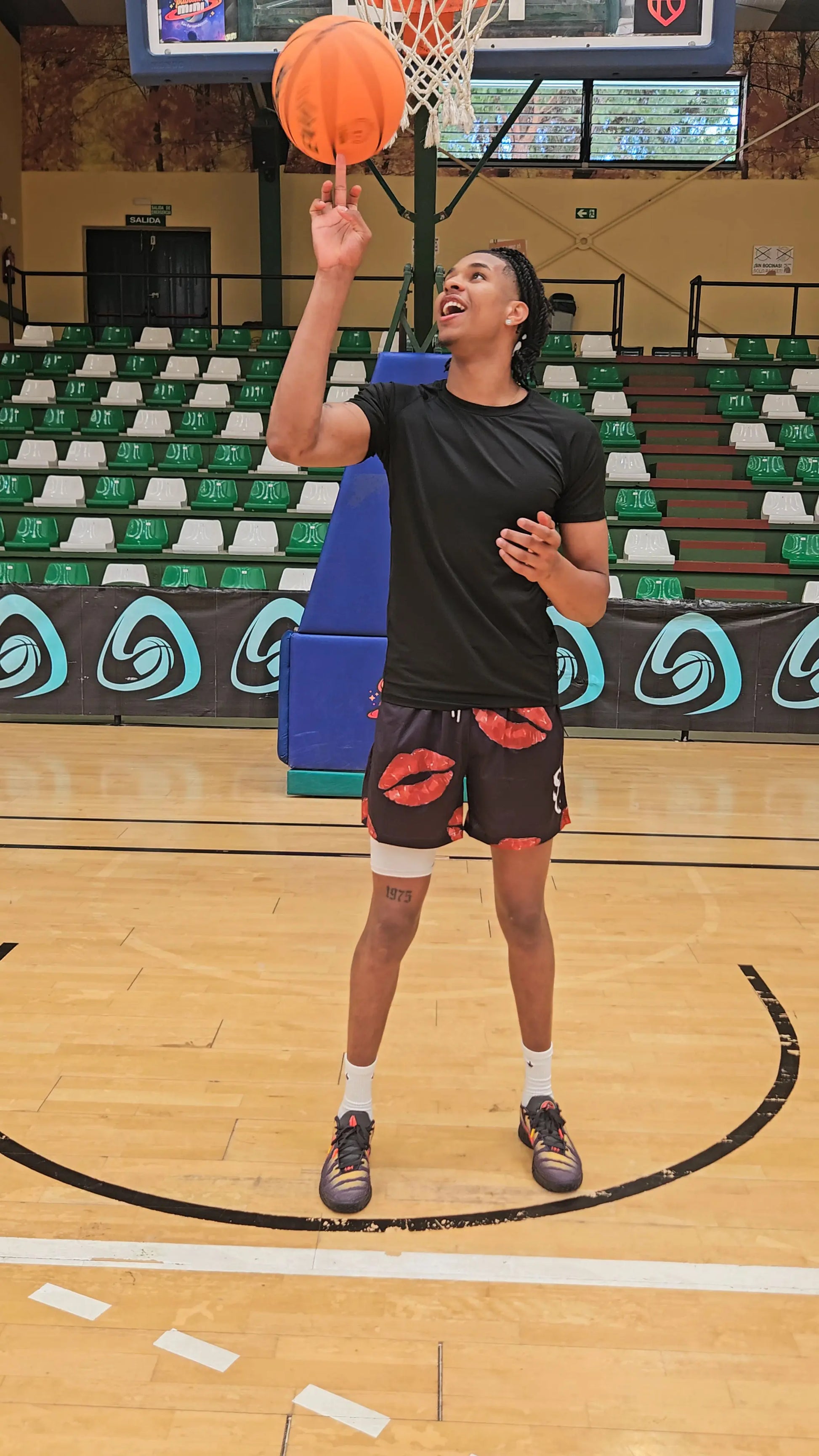 Basketball player spinning a ball on his finger while wearing black basketball shorts with a red lip/kiss print on a basketball court
