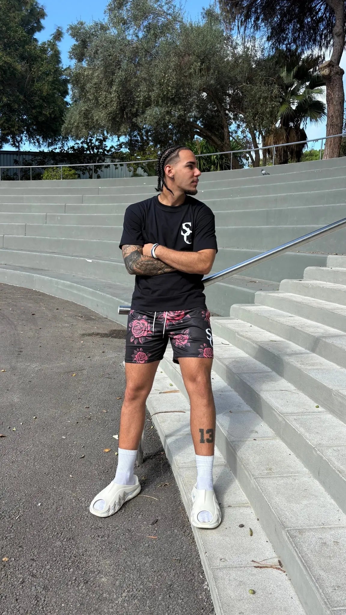 A model standing with arms crossed on outdoor steps, wearing black basketball shorts with a red rose pattern and a matching black t-shirt