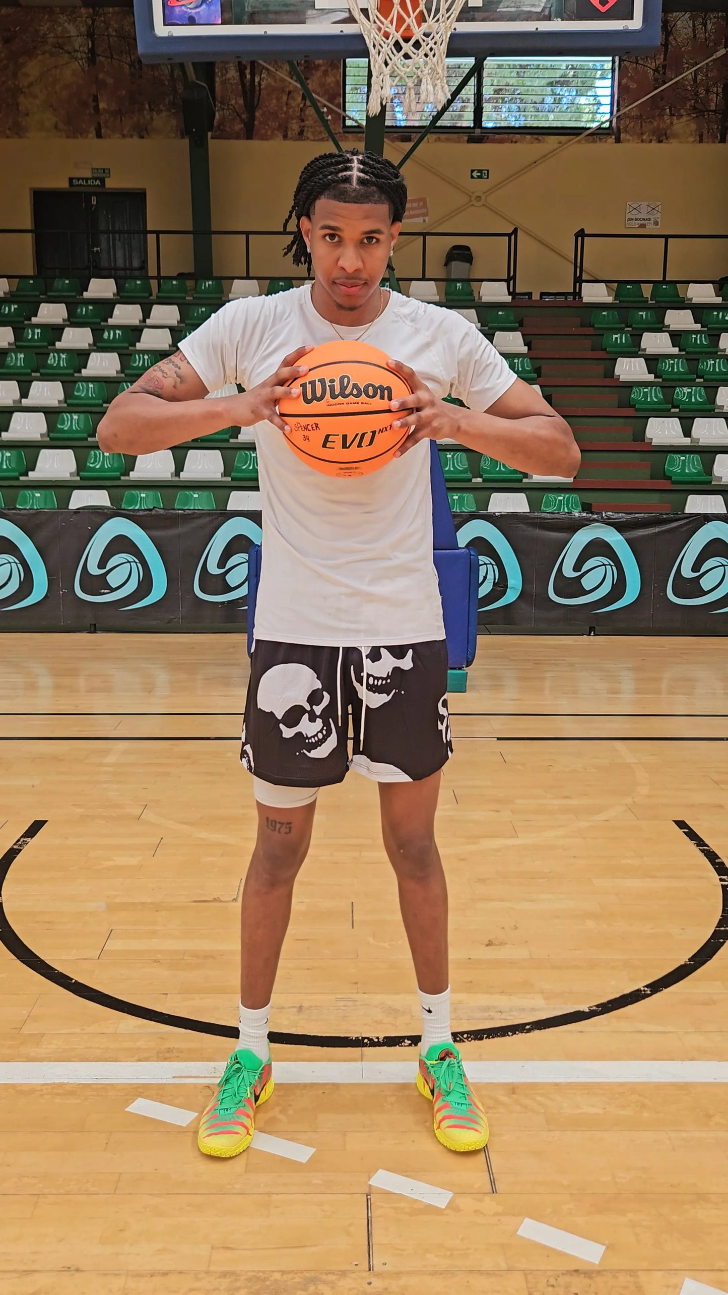 Basketball player standing on a court holding a basketball, wearing black basketball shorts with a white skull pattern
