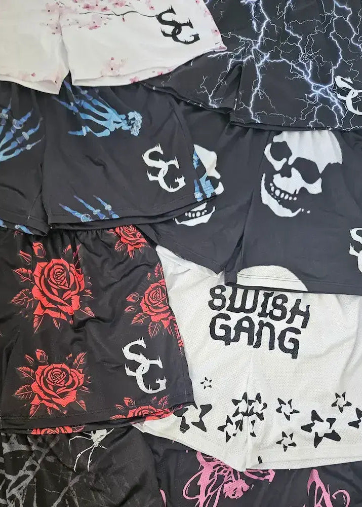 Flat lay product shot of multiple Swish Gang basketball shorts featuring various prints including cherry blossoms, lightning, skeleton hands, skulls, roses, and star graphics with SG logos.