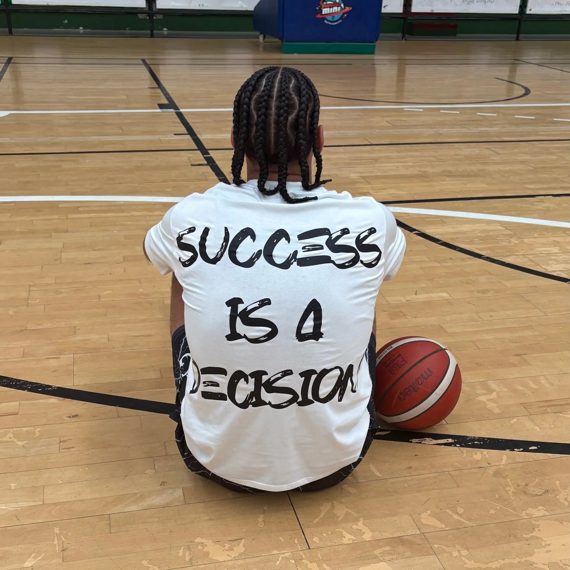 Back view of a basketball player sitting on a basketball court wearing a white SwishGang t-shirt with the bold "SUCCESS IS A DECISION" graphic