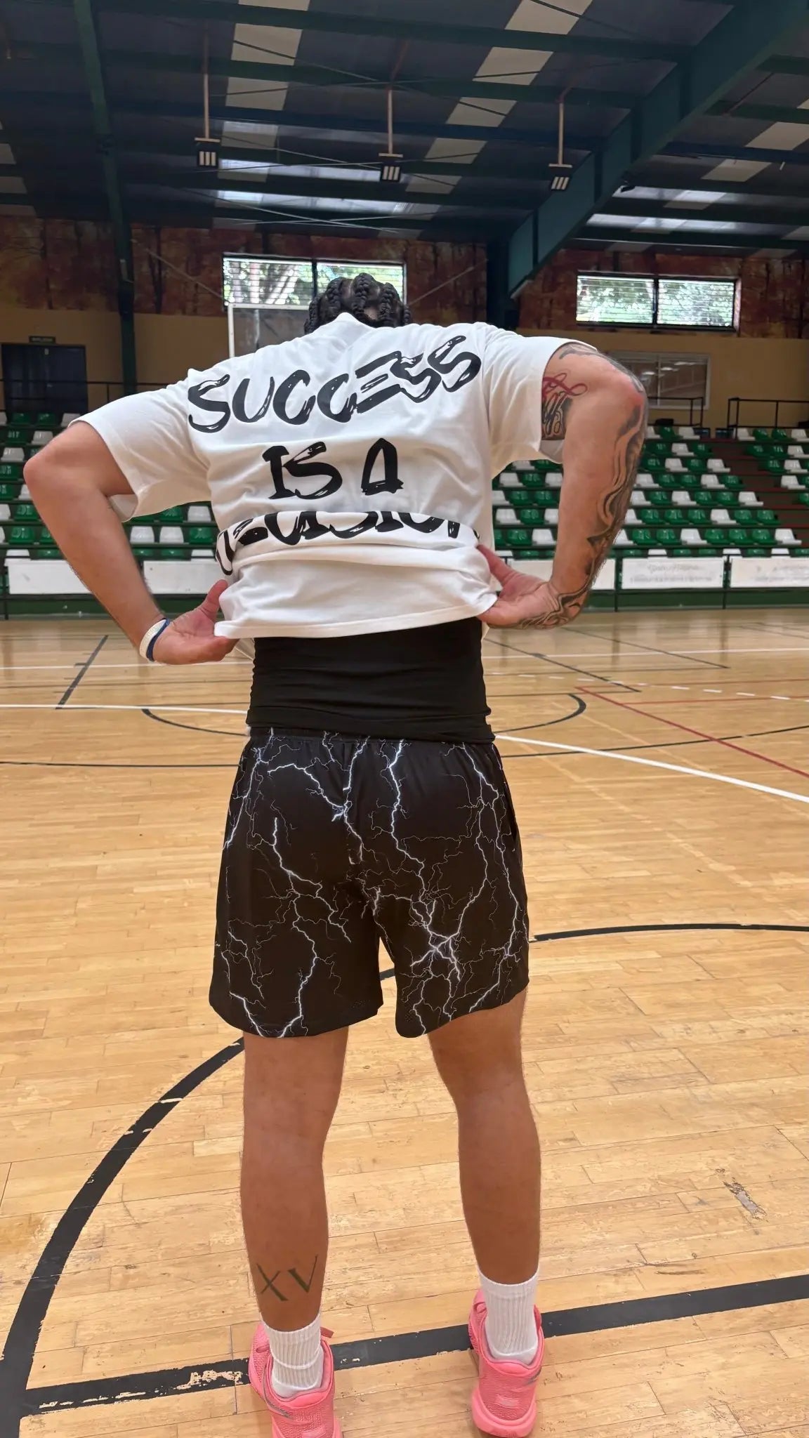 A basketball player on a court lifting the back of a white "SUCCESS IS A DECISION" t-shirt to reveal a black base layer and lightning-print shorts
