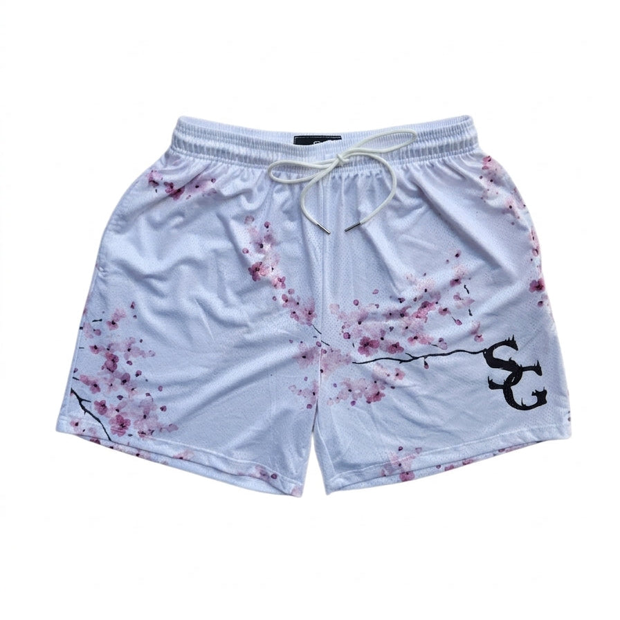 Studio product shot of white basketball shorts with a pink cherry blossom branch print and black logo on the left leg